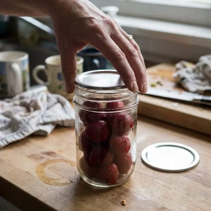 A hand placing a lid on a mason jar filled with strawberries